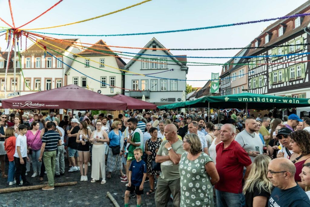 Eine große Menschenmenge versammelt sich beim Johannisfest 2025 auf dem Stadtplatz von Groß-Umstadt, umgeben von Fachwerkhäusern, Sonnenschirmen und bunten Luftschlangen. Menschen aller Altersgruppen unterhalten sich und genießen die lebhafte Festatmosphäre.