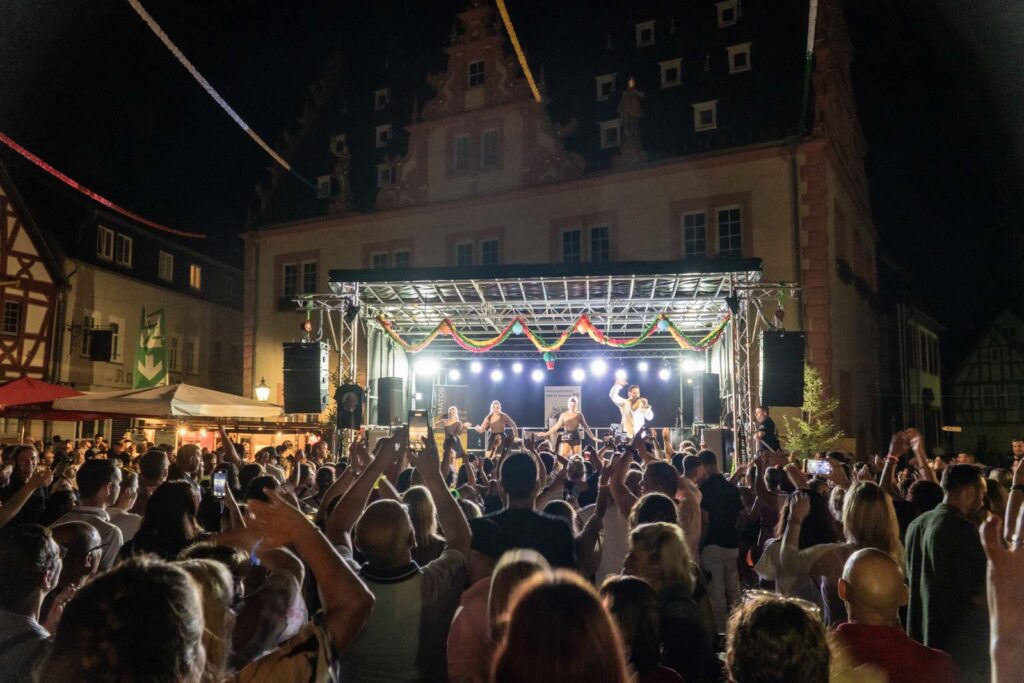 Eine große Menschenmenge versammelt sich nachts vor einer Freilichtbühne auf dem Johannisfest 2025 in Groß-Umstadt, wo eine Live-Band auftritt, bunte Lichter leuchten und historische Gebäude im Hintergrund zu sehen sind. Viele Menschen haben ihre Hände erhoben und genießen die Musik.