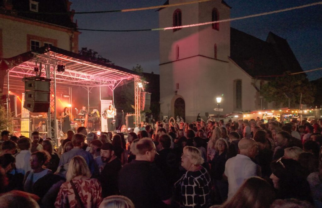 Eine große Menschenmenge versammelt sich bei einem nächtlichen Konzert im Freien während des Johannisfestes 2025 in Groß-Umstadt. Eine Band spielt auf einer beleuchteten Bühne vor einer Kirche, während Essensstände und festliche Lichter eine lebendige São-João-Atmosphäre schaffen.