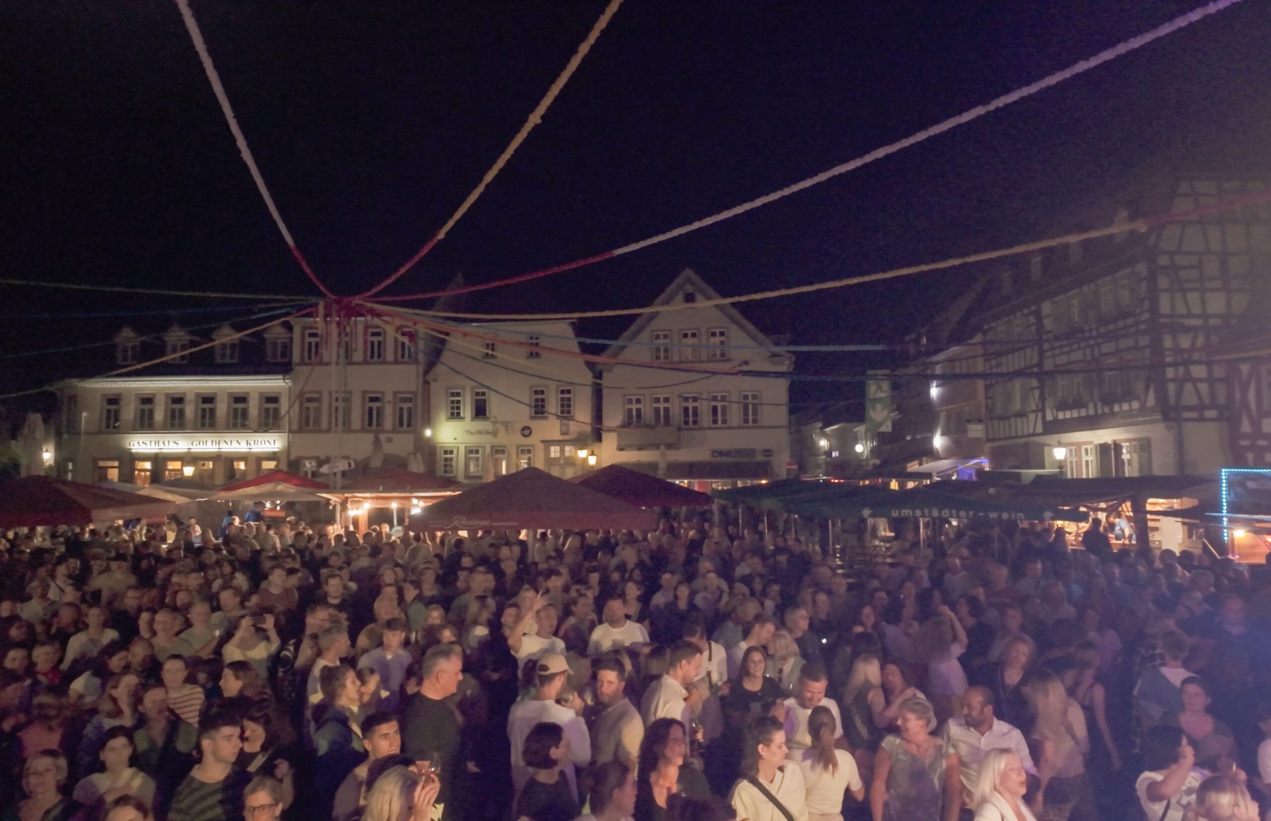 Eine große Menschenmenge versammelt sich auf dem Johannisfest 2025 auf dem Marktplatz von Groß-Umstadt, umgeben von historischen Gebäuden, beleuchteten Ständen und Lichterketten über dem Kopf. Die Atmosphäre wirkt lebendig und festlich und erinnert an den Geist der São-João-Feiern.