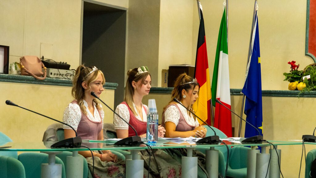 Três mulheres em traje tradicional no púlpito, diante das bandeiras de Itália e da Europa, num encontro de geminação em Dicomano