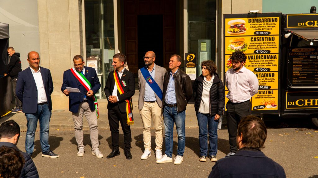 Eine Gruppe von sieben Personen steht in einer Reihe im Freien in der Nähe eines Foodtruck-Menüs auf der Fiera d'Ottobre, mit drei Männern, die Schärpen tragen. Ein Mann liest aus einer Zeitung vor, während die anderen auf die Veranstaltung "Partnerstadt Dicomano" hinweisen.
