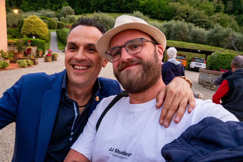 Zwei Männer posieren für ein fröhliches Selfie im Freien während ihrer Verschwisterungsreise. Der eine trägt eine blaue Jacke, der andere ein weißes T-Shirt und einen Hut. Im Hintergrund sind üppiges Grün, eine Einfahrt und geparkte Autos zu sehen.