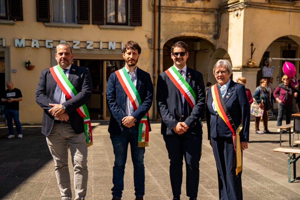 Vier Personen, die Schärpen in den Farben der italienischen Flagge tragen, stehen im Freien auf einem sonnigen Platz zusammen, wahrscheinlich während einer Verschwisterungsreise, wobei im Hintergrund Gebäude und mehrere andere Personen zu sehen sind.