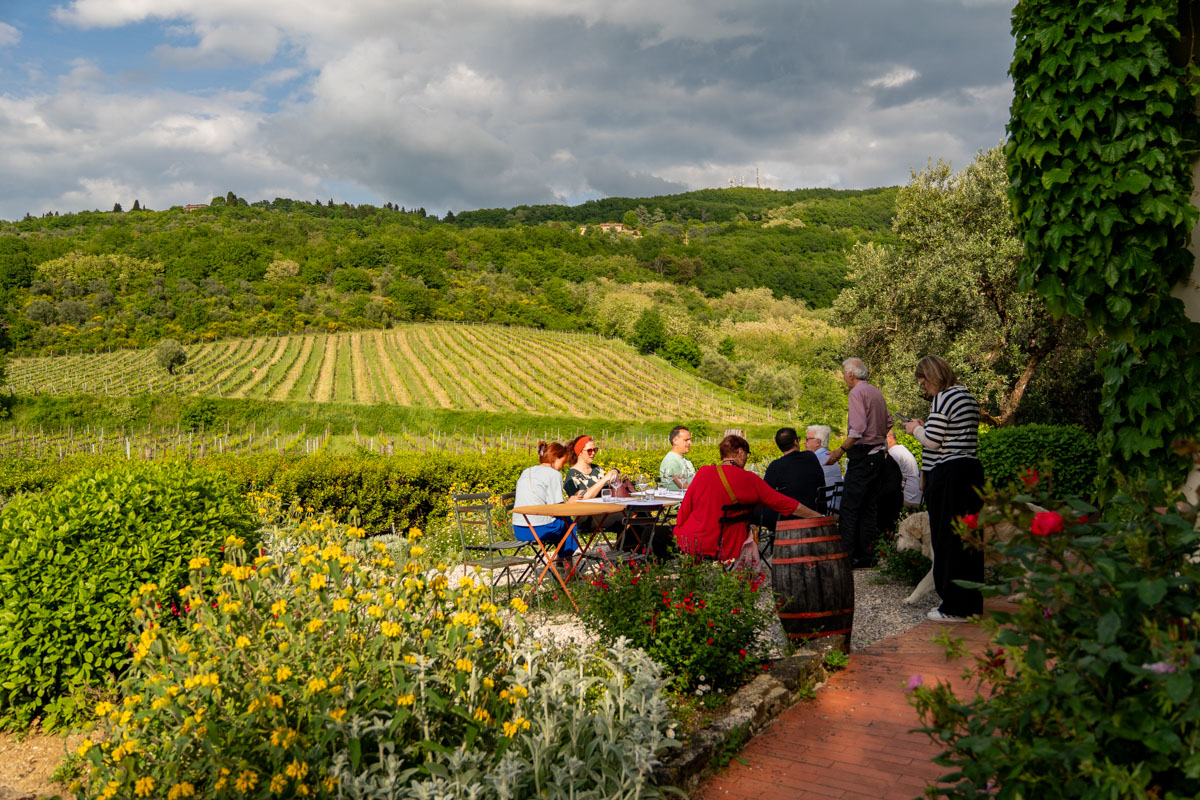 Eine Gruppe von Menschen sitzt an Tischen im Freien und genießt Essen und Gespräche während einer Verschwisterungsreise, umgeben von üppigen Gärten und gelben Blumen, mit grünen Weinbergen und sanften Hügeln im Hintergrund unter einem teilweise bewölkten Himmel.