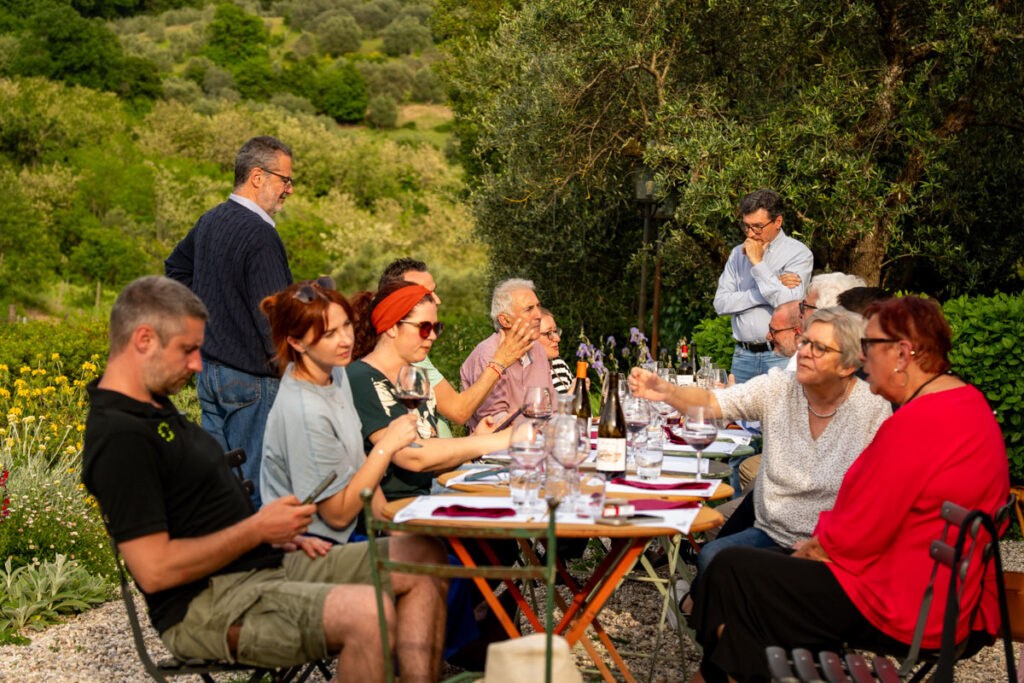 Eine Gruppe von Menschen unterschiedlichen Alters versammelt sich um einen langen Tisch im Freien und genießt eine Mahlzeit während einer Verschwisterungsreise in einer üppigen Gartenlandschaft. Einige unterhalten sich, essen oder schauen inmitten der Bäume und des Grüns auf ihr Handy.