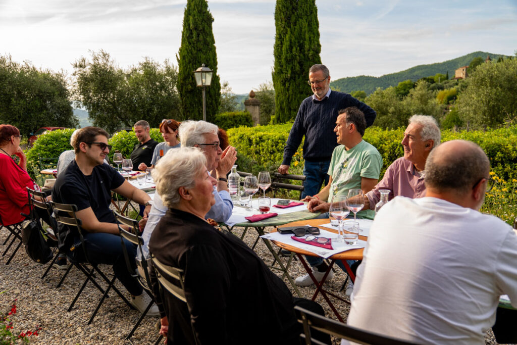 Eine Gruppe von Menschen genießt eine Verschwisterungsreise, isst und unterhält sich an Tischen im Freien, umgeben von Grün und Hügeln. Einige lachen, während andere einem Mann zuhören, der spricht. Weingläser und Teller glänzen im Sonnenlicht.