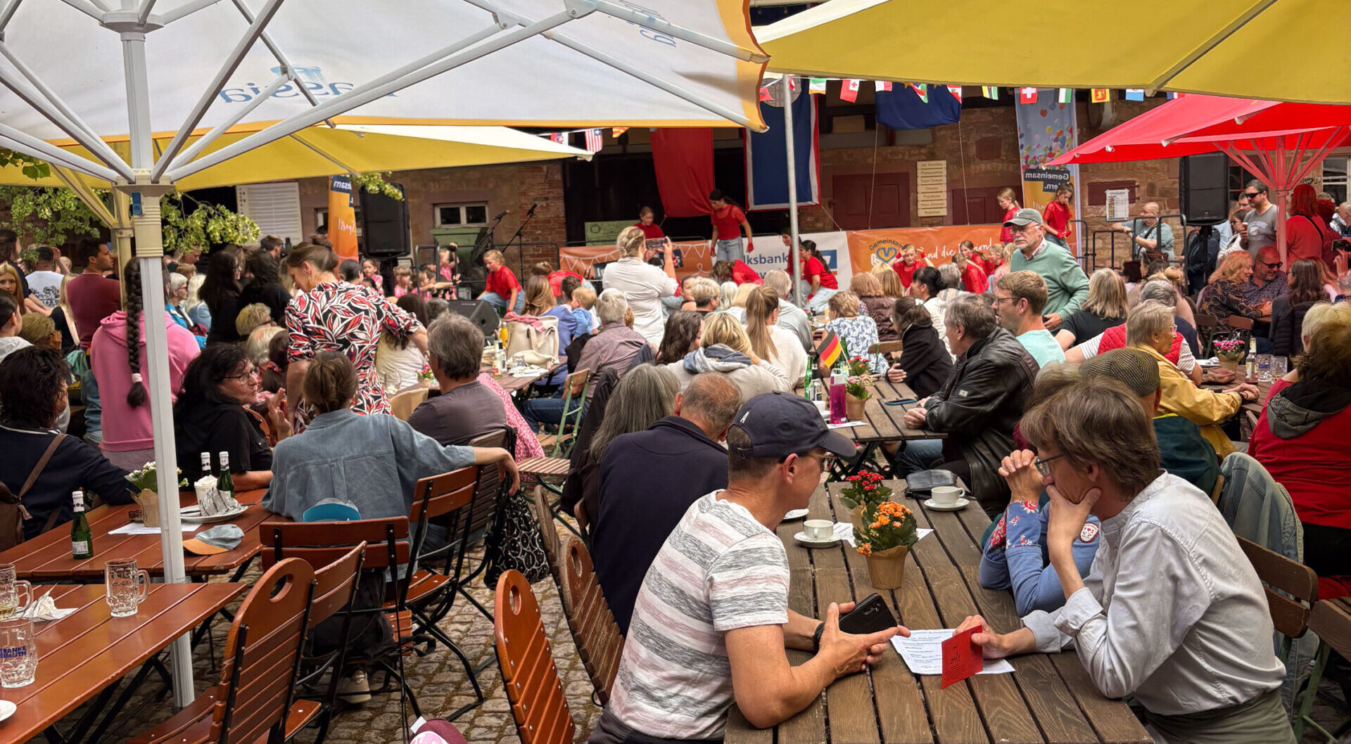 Während des Internationalen Sommerfestes versammeln sich die Menschen an Holztischen im Freien unter bunten Sonnenschirmen in einem lebhaften Café im Innenhof. Viele essen, trinken und unterhalten sich, während Dekorationen und eine vom Verschwisterungskomitee organisierte Band für Stimmung sorgen.