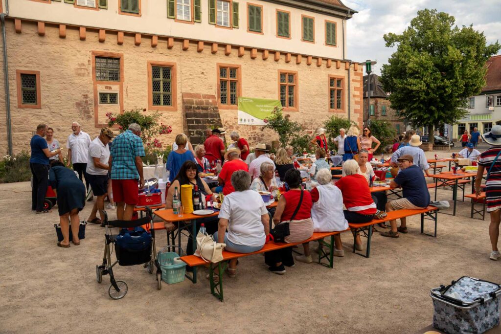In Groß-Umstadt versammeln sich Menschen an Picknicktischen im Freien, essen und unterhalten sich in der Nähe eines alten Steingebäudes. Auf den Tischen stehen Kühlboxen, Essen und Getränke – das erinnert an ein ungezwungenes „Dîner en bleu blanc rouge“. Einige Leute stehen oder gehen in der Nähe umher und tragen so zur festlichen Atmosphäre der Szene bei.