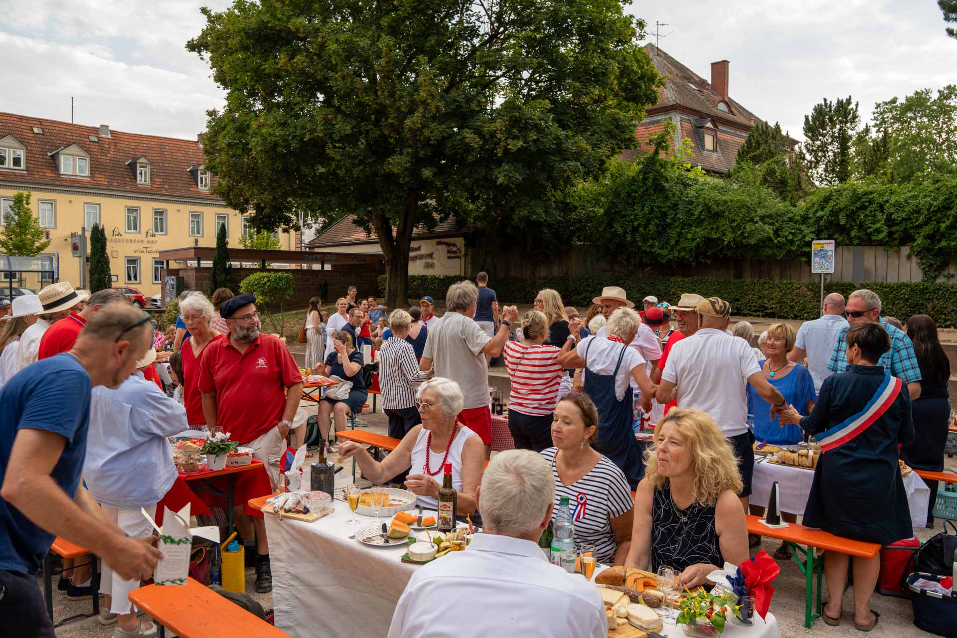Bei einem „Dîner en bleu blanc rouge“ in Groß-Umstadt versammeln sich die Menschen im Freien um lange Tische mit Speisen und feiern gemeinsam. Einige tragen rote, weiße und blaue Kleidung. Im Hintergrund sind an diesem sonnigen Tag Bäume und Gebäude zu sehen, die den Rahmen für einen wahrhaft grandiosen Abend bilden.