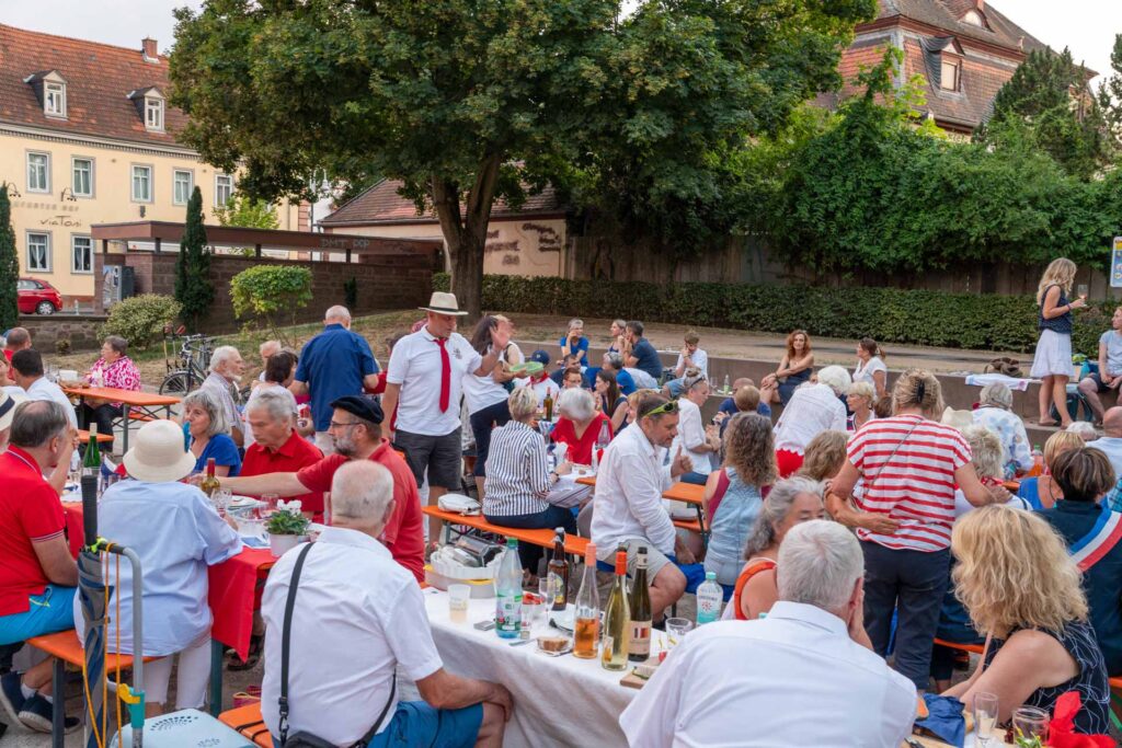 Eine große Gruppe von Menschen versammelt sich im Freien an langen Tischen und genießt Speisen und Getränke bei einer festlichen Veranstaltung namens „Dîner en bleu blanc rouge“ in Groß-Umstadt. Viele tragen Sommerkleidung und Hüte, was zur lebhaften Atmosphäre beiträgt. Im Hintergrund sind Bäume und Gebäude zu sehen.