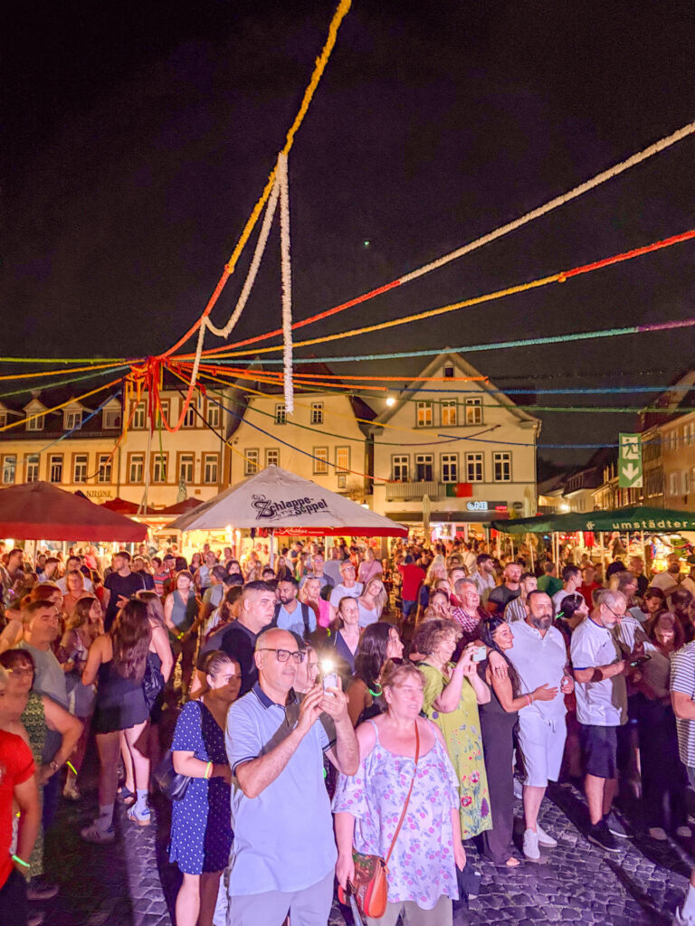 Während des Johannisfests in Groß-Umstadt versammelt sich nachts eine große Menschenmenge auf dem festlich geschmückten Stadtplatz, der mit bunten Luftschlangen verziert ist. Die Stimmung ist ausgelassen, während die Menschen lächeln und Fotos machen – inmitten beleuchteter Gebäude und lebhafter Wochenendmarktstände.