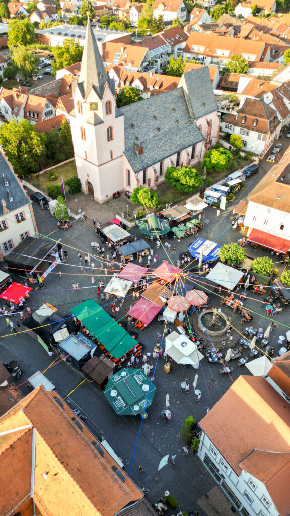 Vista aérea da praça do mercado durante a festa de São João, com a igreja, bancas e um mastro decorado com fitas coloridas, em redor do qual as pessoas se reúnem.