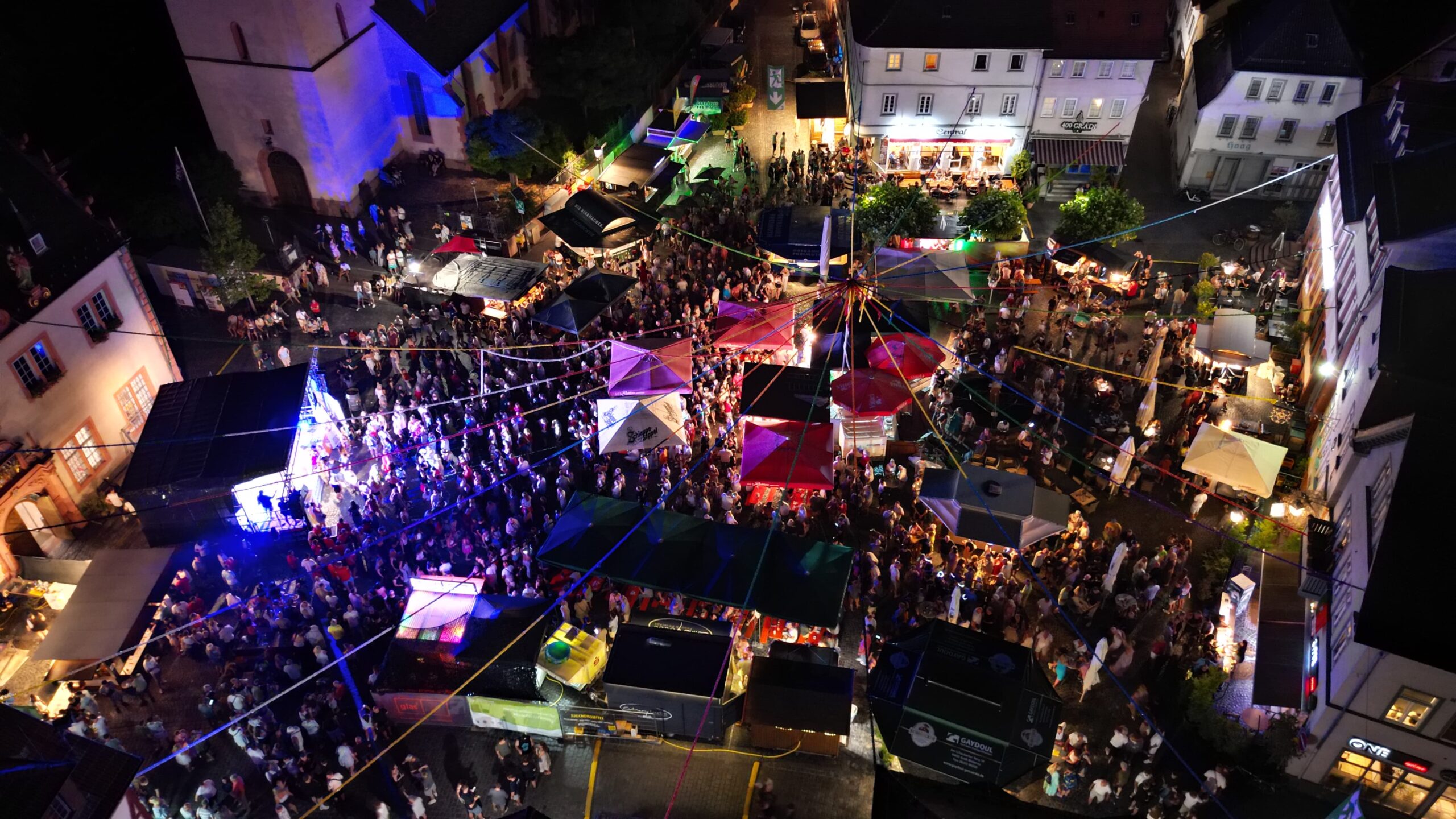Luftaufnahme des Johannisfests bei Nacht mit bunten Zelten, Imbissständen, Lichterketten und einer großen Menschenmenge, die sich zwischen den Gebäuden versammelt hat und für eine lebhafte Feststimmung sorgt.