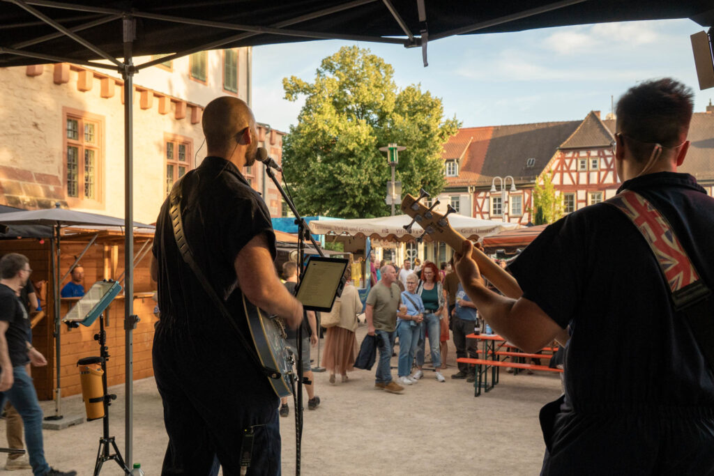 Bei einer Open-Air-Veranstaltung auf einem malerischen Stadtplatz, umgeben von historischen Gebäuden und Menschen, die sich beim lebhaften Verschwisterungsfest Groß-Umstadt unterhalten, treten zwei Musiker auf der Bühne vor einem Publikum auf.